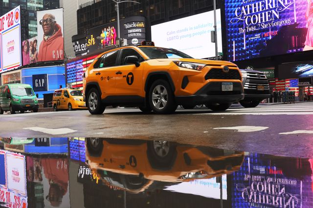 A yellow cab SUV in Times Square, with billboards seen in the background. The taxi's reflection can be seen in a puddle in the foreground of the photo